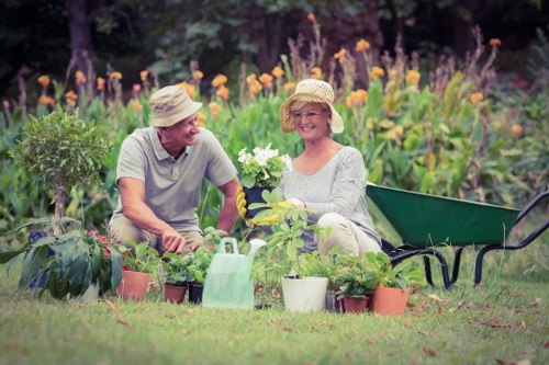 A smiling middle-aged man and woman sit in a well-maintained garden, surrounded by various potted plants, gardening tools, and a watering can. The woman holds a white flowering plant, while the man appears engaged in gardening activities. Behind them, there is a lush flower bed with tall purple and orange flowers, and the area is bordered by trees and greenery. The scene takes place outdoors on a bright, sunny day, with natural lighting highlighting the vibrant colors of the flowers and plants, suggesting a peaceful, landscaped garden space typical of residential outdoor areas in Canary Wharf. This setting reflects professional gardening and landscaping work, with a focus on plant care and outdoor maintenance, supported by local services such as hedge trimming and lawn care providers like Hedge Trimming Canary Wharf.