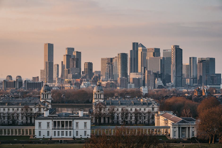 A wide view of a city skyline during sunset, featuring a modern cluster of high-rise office buildings and residential towers, with a historic white mansion in the foreground. The mansion has classical