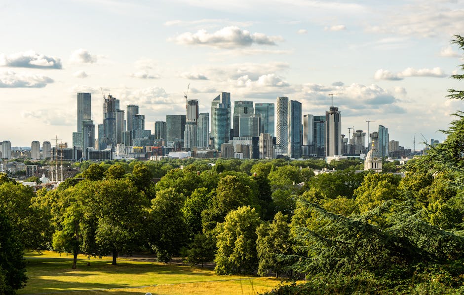 A wide view of a city skyline during sunset, featuring a modern cluster of high-rise office buildings and residential towers, with a historic white mansion in the foreground. The mansion has classical