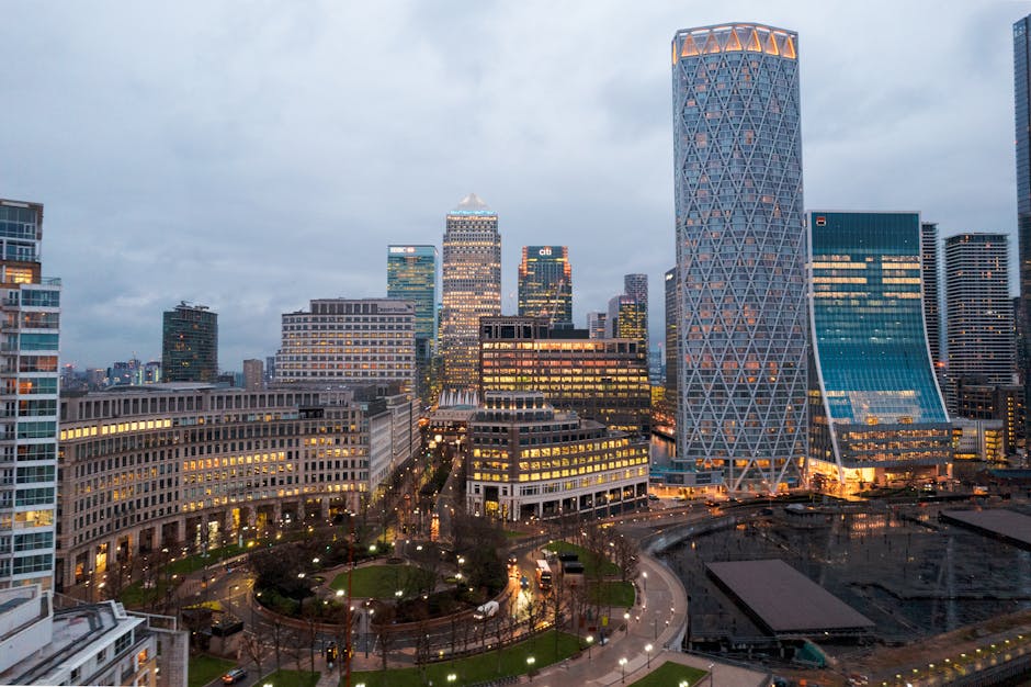 A panoramic view of the Canary Wharf skyline during the early evening, featuring tall, modern skyscrapers with glass facades reflecting the last light of the day. In the foreground, a well-maintained 