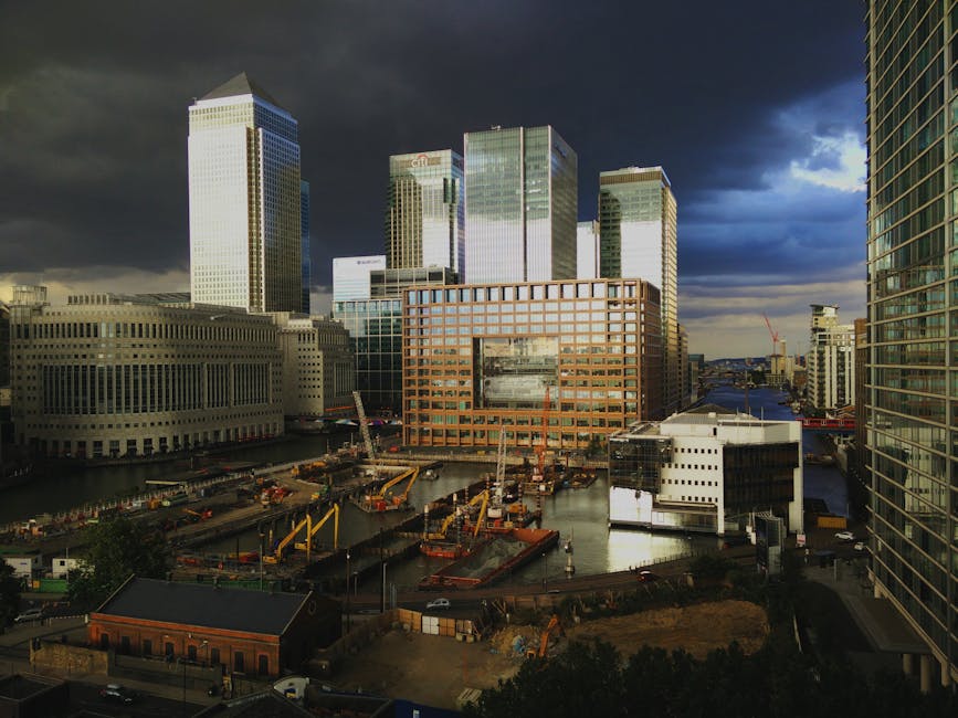 A panoramic view of the Canary Wharf skyline during the early evening, featuring tall, modern skyscrapers with glass facades reflecting the last light of the day. In the foreground, a well-maintained 