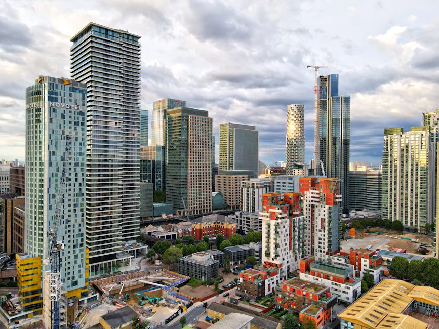 A cityscape featuring modern skyscrapers with glass facades reflecting the cloudy sky, interspersed with mid-rise buildings, some under construction with cranes visible. In the foreground, there are s